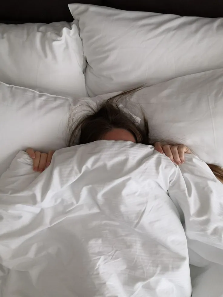 A young person with long brown hair lying in bed, mostly covered by white blankets