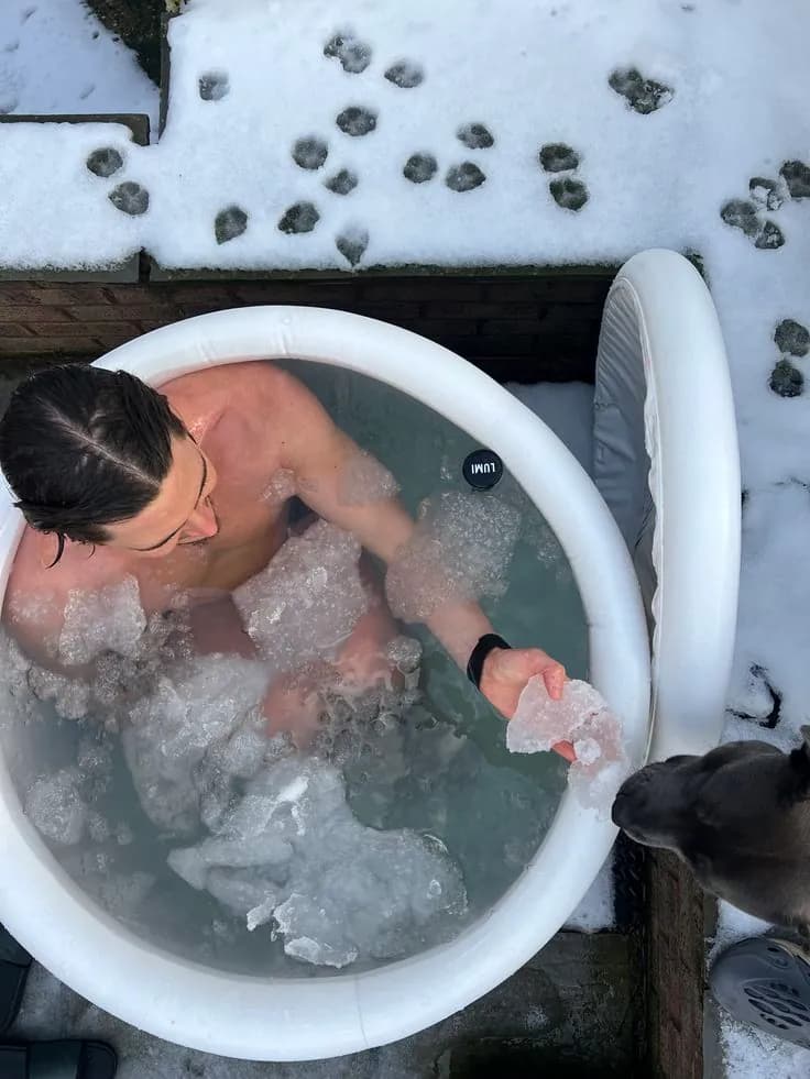 Man in an outdoor hot tub holding ice near a dog, with snow around the tub