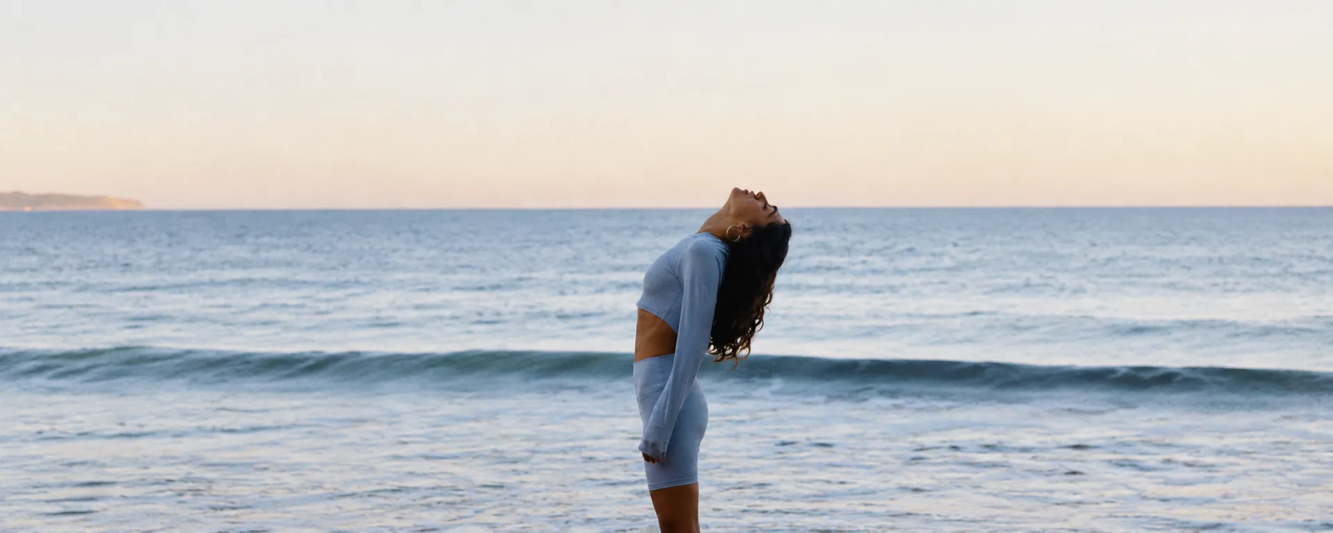 A woman in light blue activewear standing in the shallows at dawn, head tilted back to the sky
