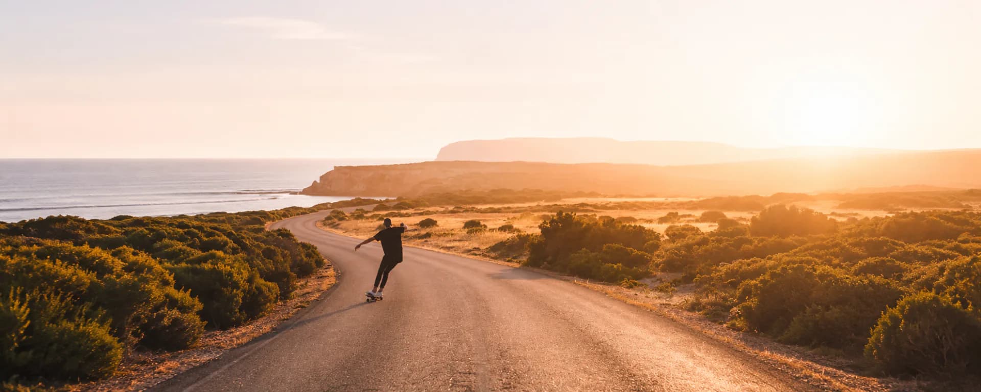 A skateboarder riding a winding coastal road at golden hour