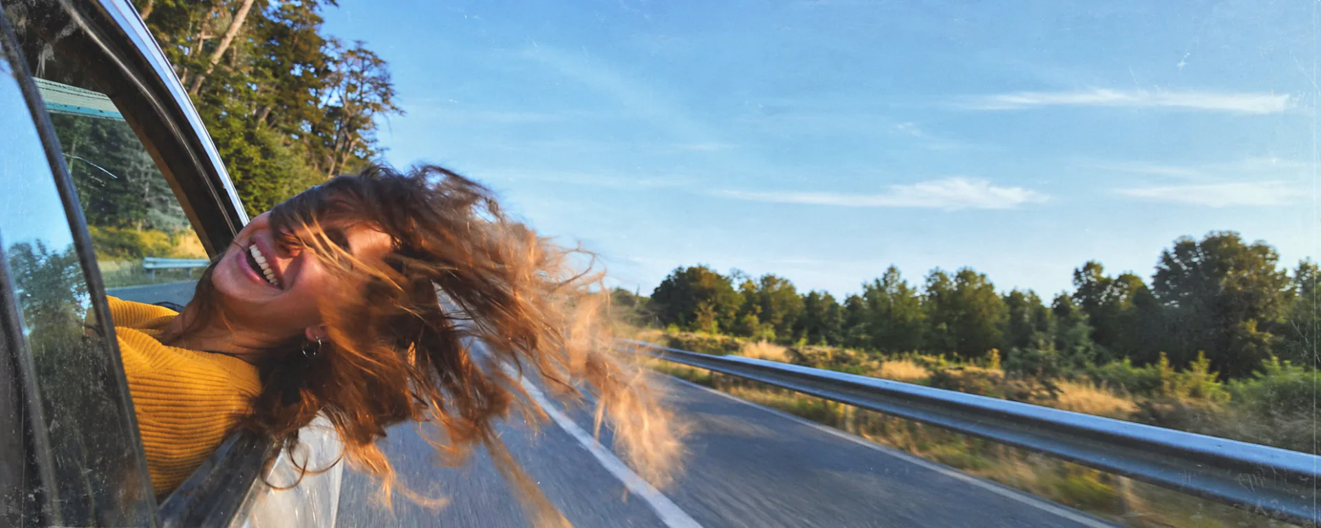 A woman leaning out of a moving car window, laughing, with her hair in the wind