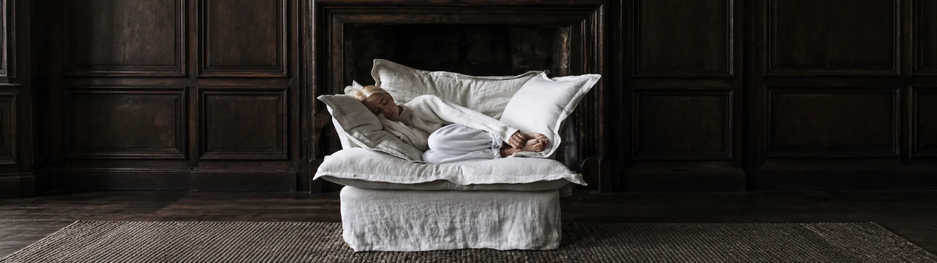 A person asleep under white linen on a daybed in a dark timber-panelled room