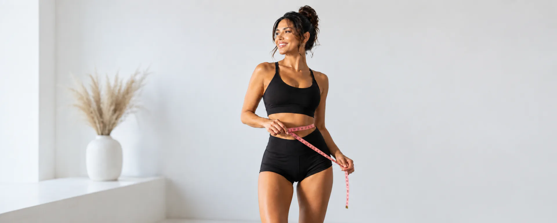 A smiling woman in black activewear holding a measuring tape around her waist in a bright white room