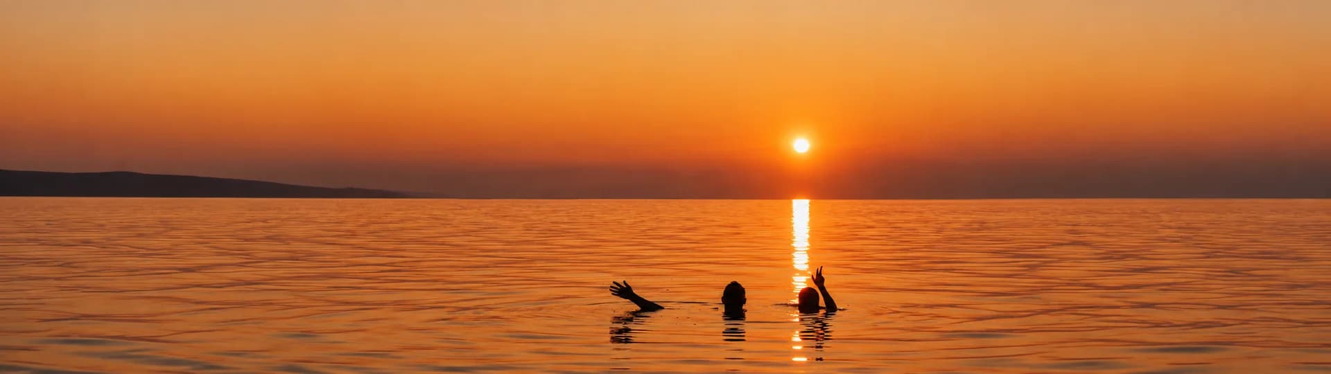 Two swimmers waving from the sea at sunset, the sun low over the horizon