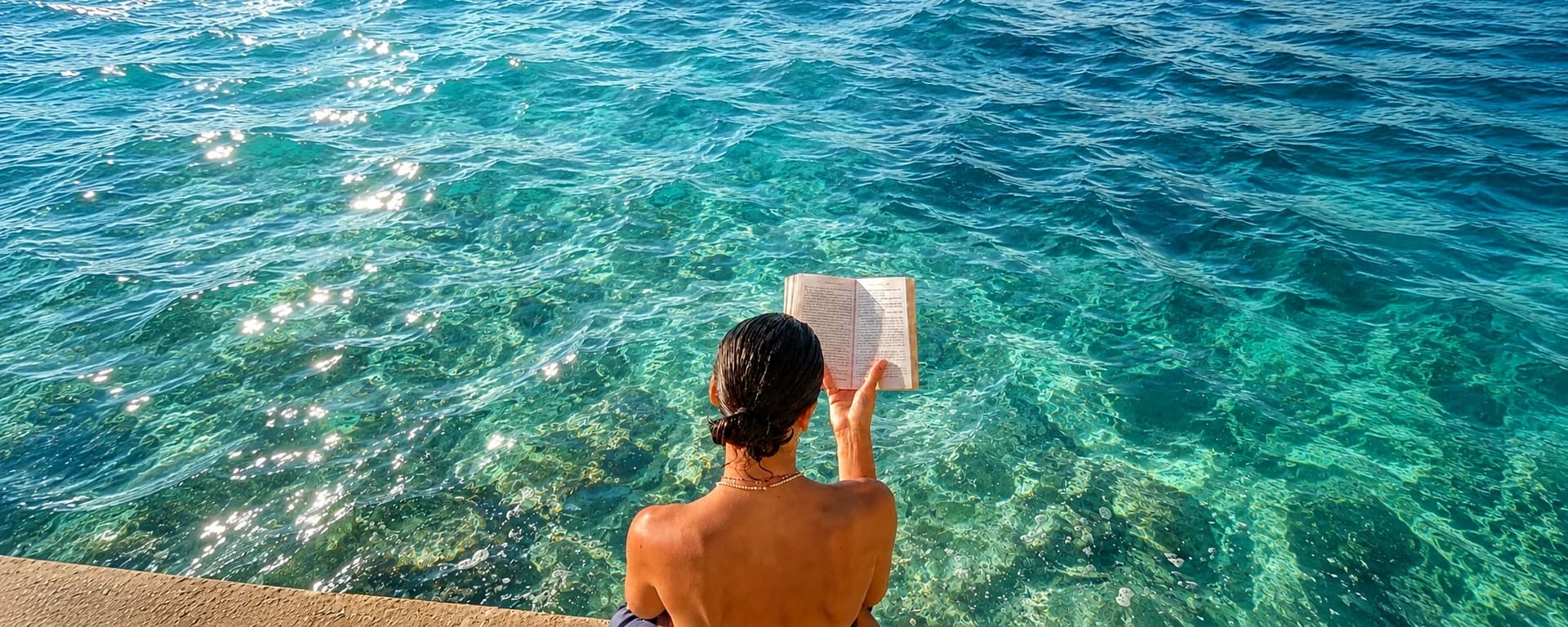 Person seen from behind reading a book on a rock ledge above clear turquoise sea